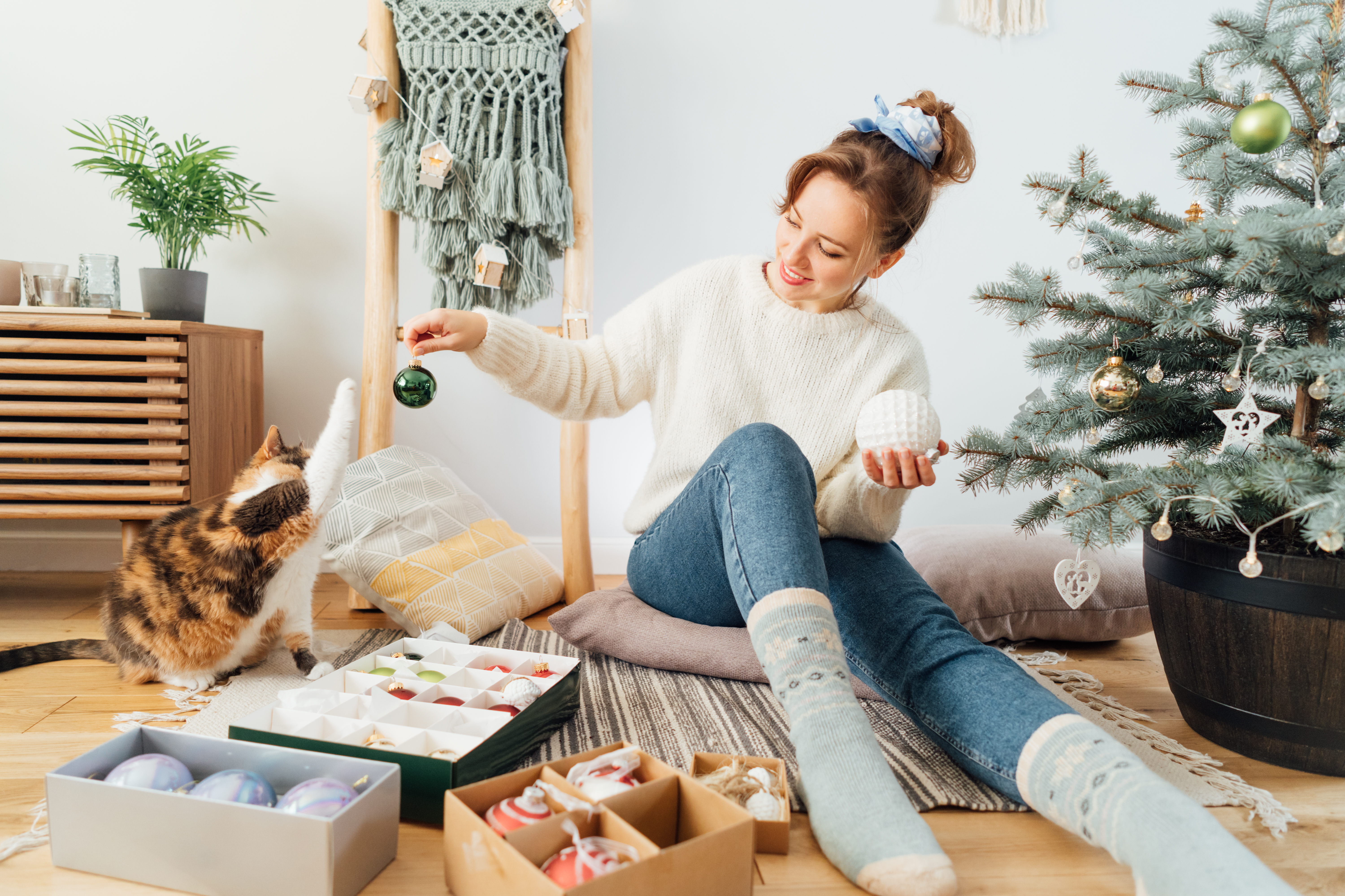 Young woman in cozy sweater decorating Christmas tree with her cat. Pet is playing with glass bubble in light modern Scandinavian interior. Winter holidays with domestic pet at home. Selective focus.